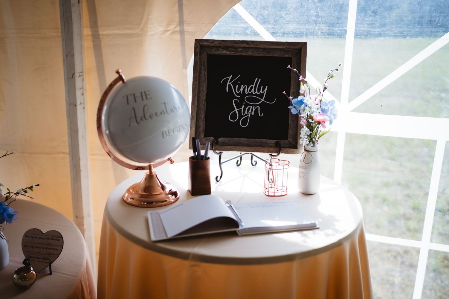 A guest book signing table at a wedding with a decorative globe, chalkboard sign, and flower vase by a window.