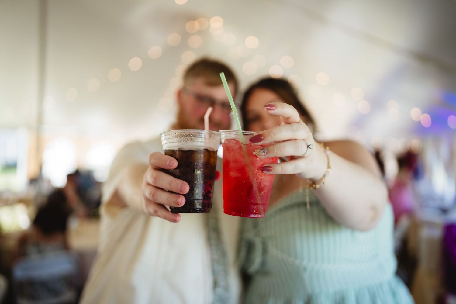 Two hands clink glasses with dark and red beverages together in celebration at an indoor event.