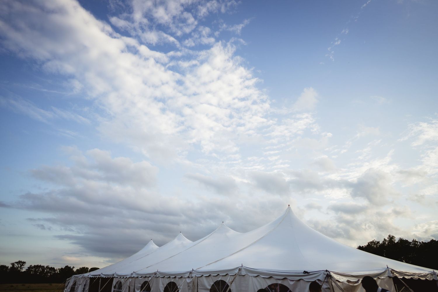 Large white outdoor event tent with sweeping peaks set against a dramatic cloudy blue sky on a sunny day.
