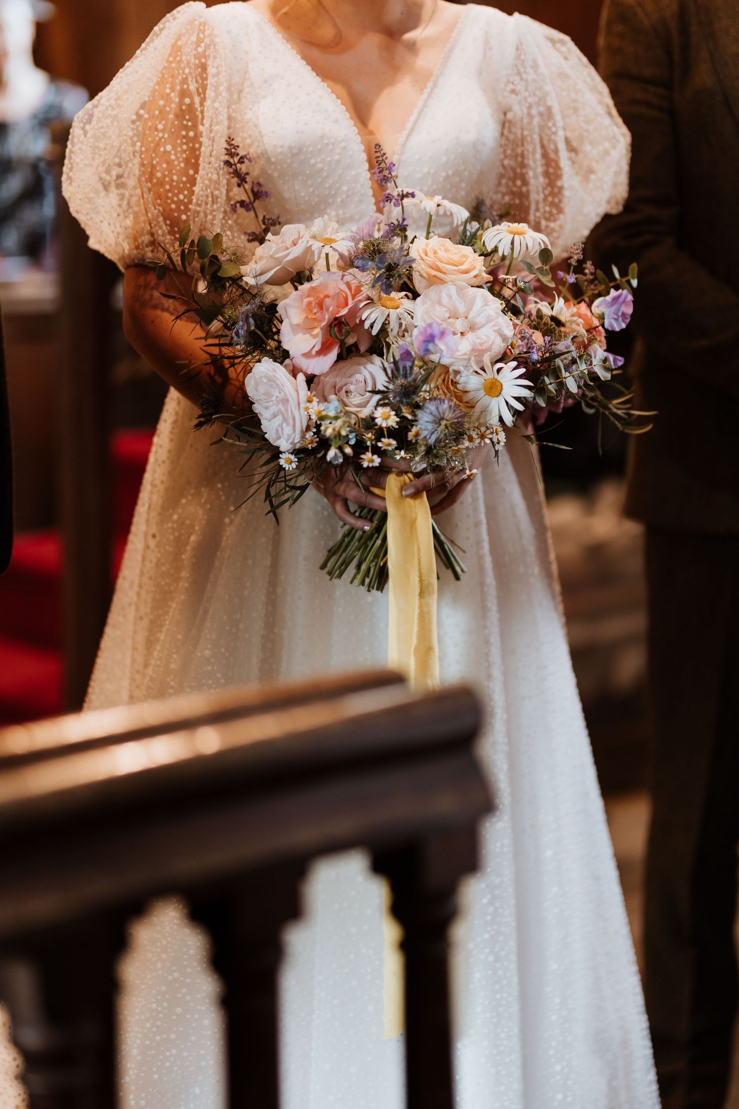 A beautiful wedding bouquet with pink and purple flowers is held against a flowing white vintage-style dress with puff sleeves.