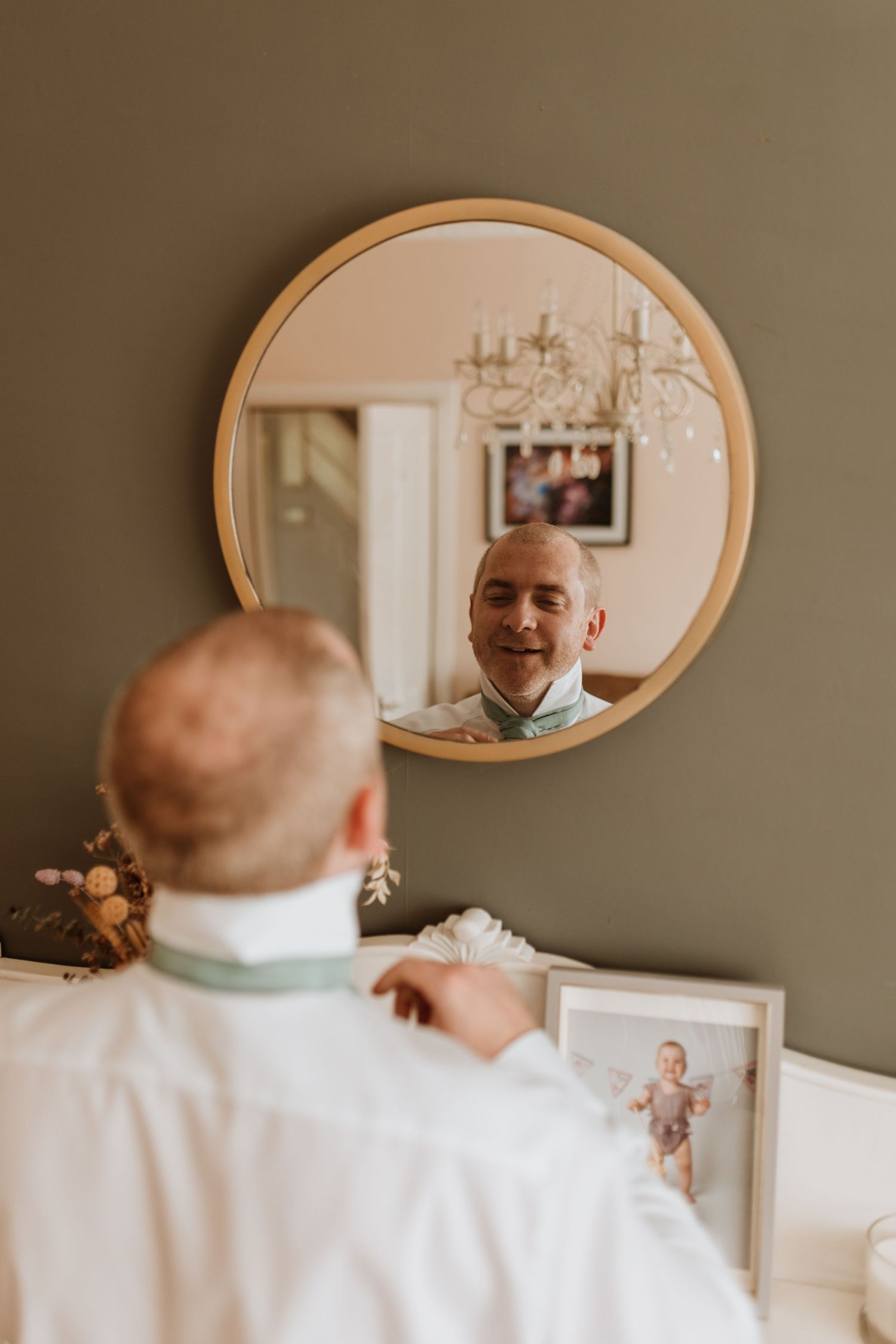 A reflection shows groom getting ready in a round gold mirror mounted on a gray wall with a chandelier visible in the background.