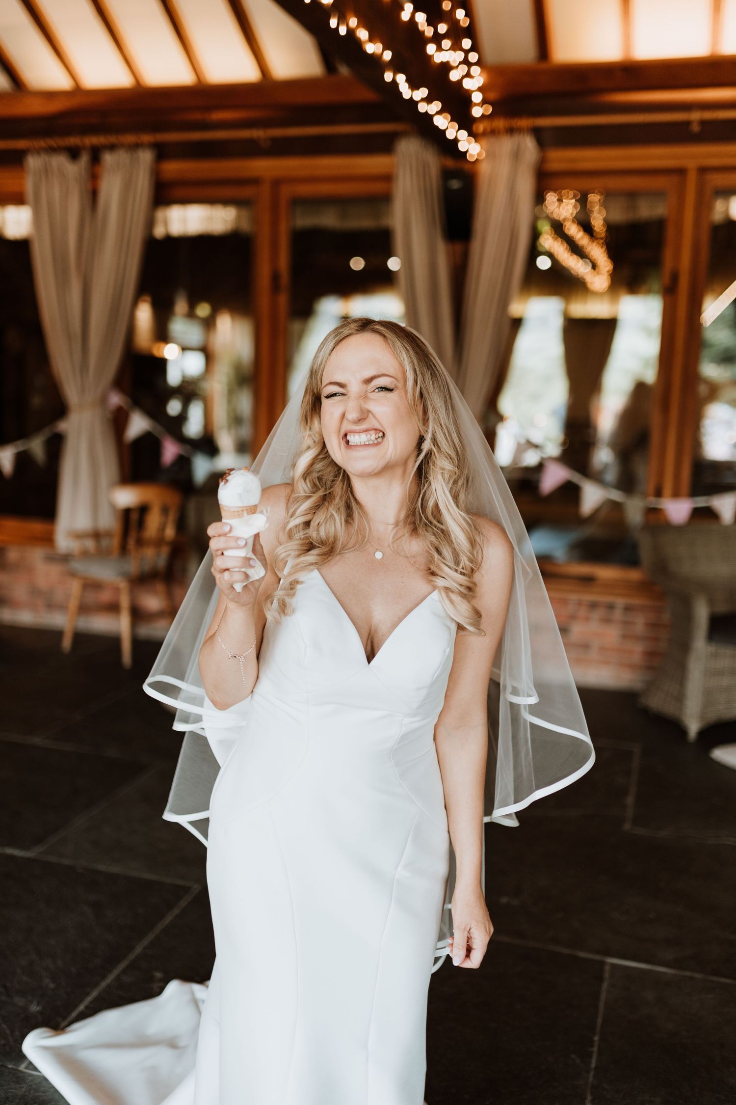 A joyful bride in a white wedding dress with a veil laughs while celebrating in a rustic indoor venue with twinkling lights.