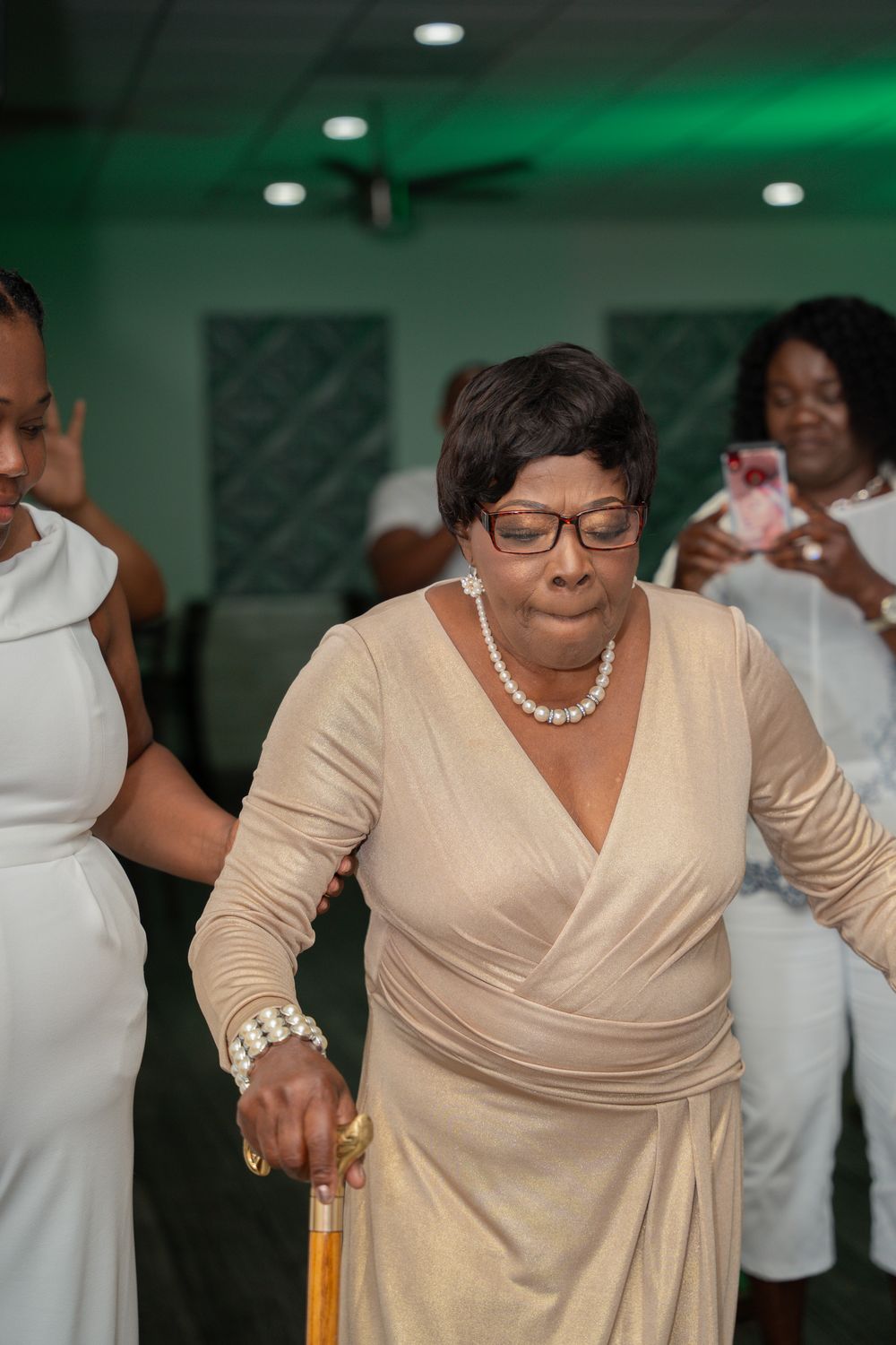 People dancing at an event with green lighting, wearing formal attire including a beige wrap dress with pearl necklace.