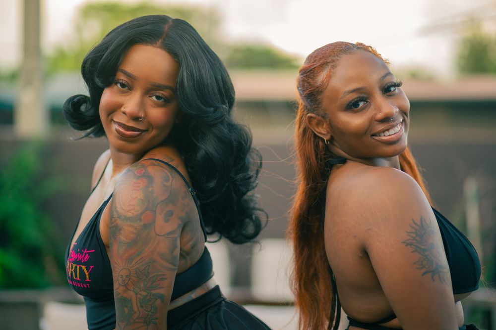 Two smiling women with tattoos pose back-to-back outdoors, one with black wavy hair and one with auburn hair.