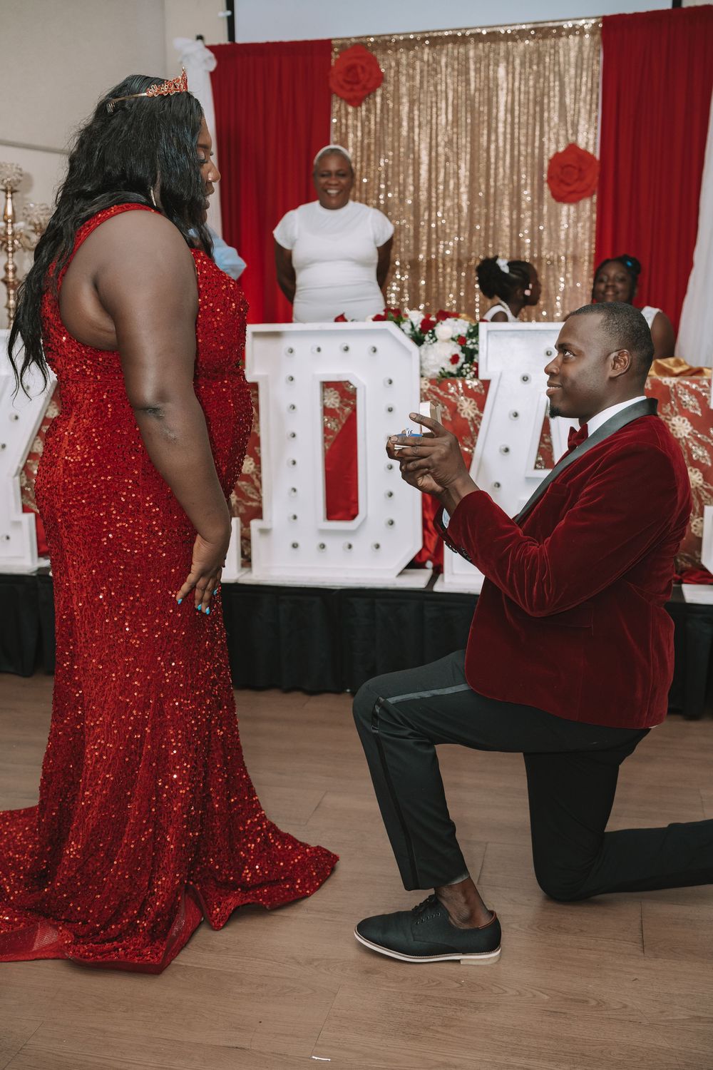 A proposal scene with someone in a red sparkly dress and another in a burgundy blazer kneeling in front of illuminated letters.