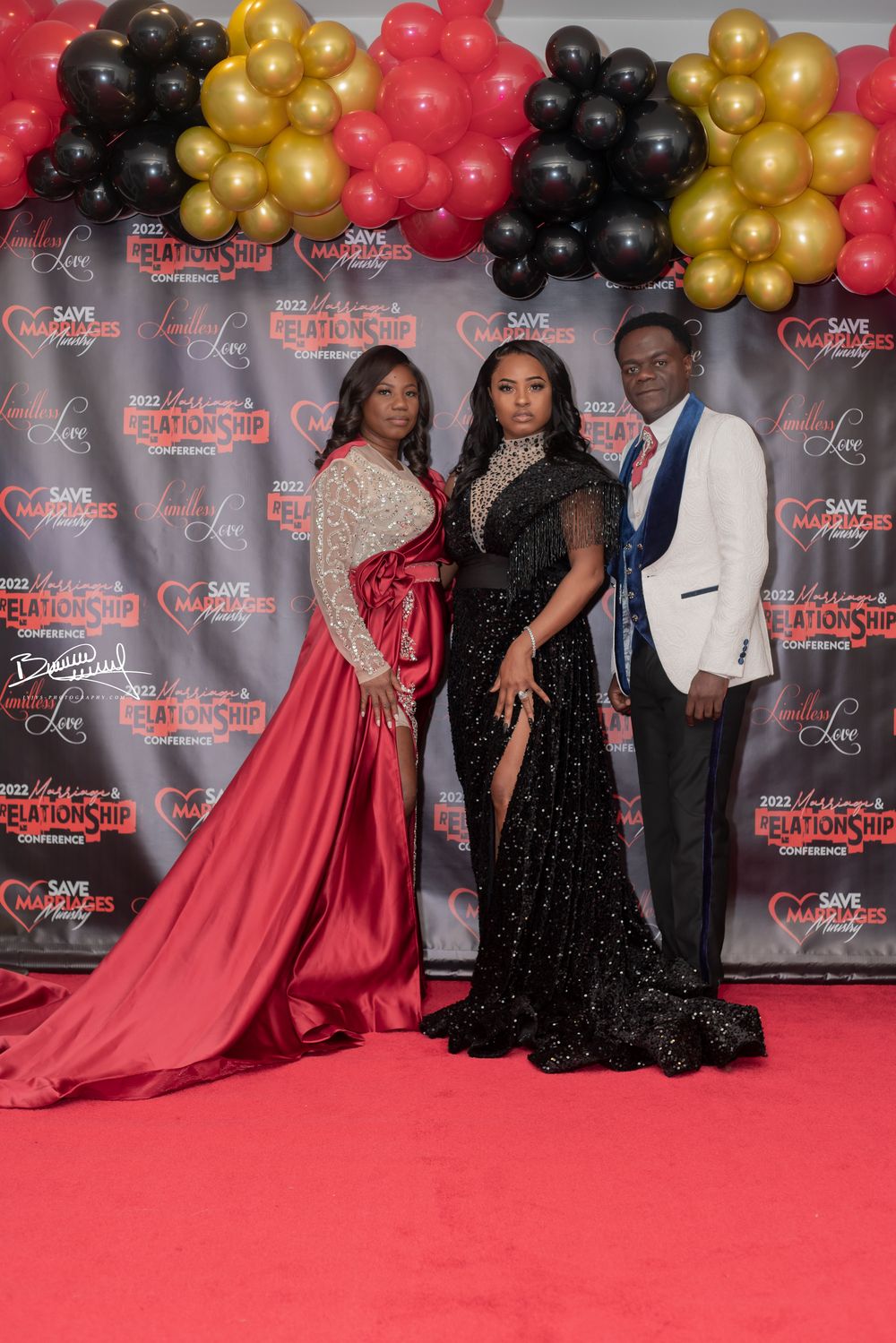 Three people pose at a formal event on a red carpet with black, gold and red balloon arch decorations in the background.