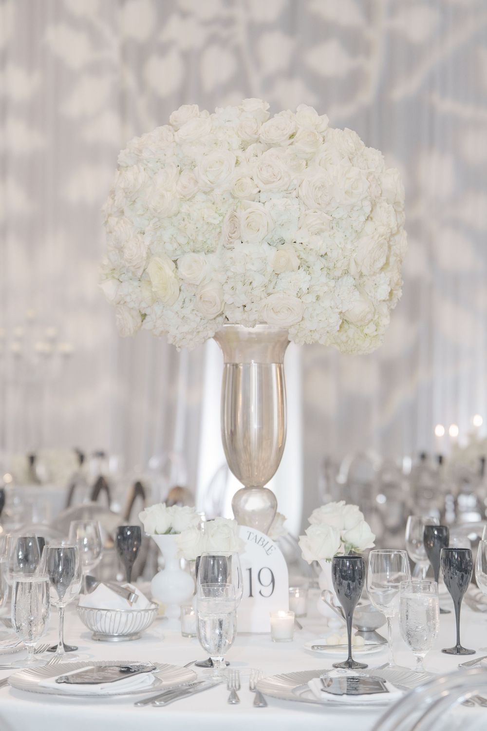 An elegant white hydrangea centerpiece in a tall silver vase adorns a formal table setting with crystal stemware.