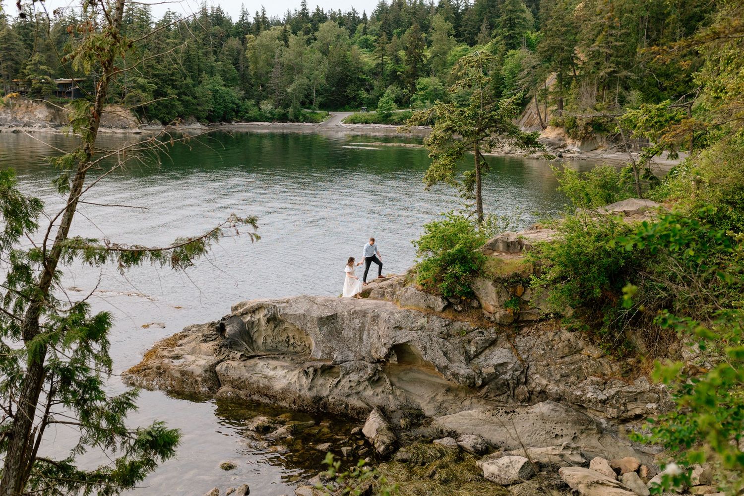 Two people stand on a rocky outcrop overlooking a serene lake surrounded by evergreen forest in the Pacific Northwest.