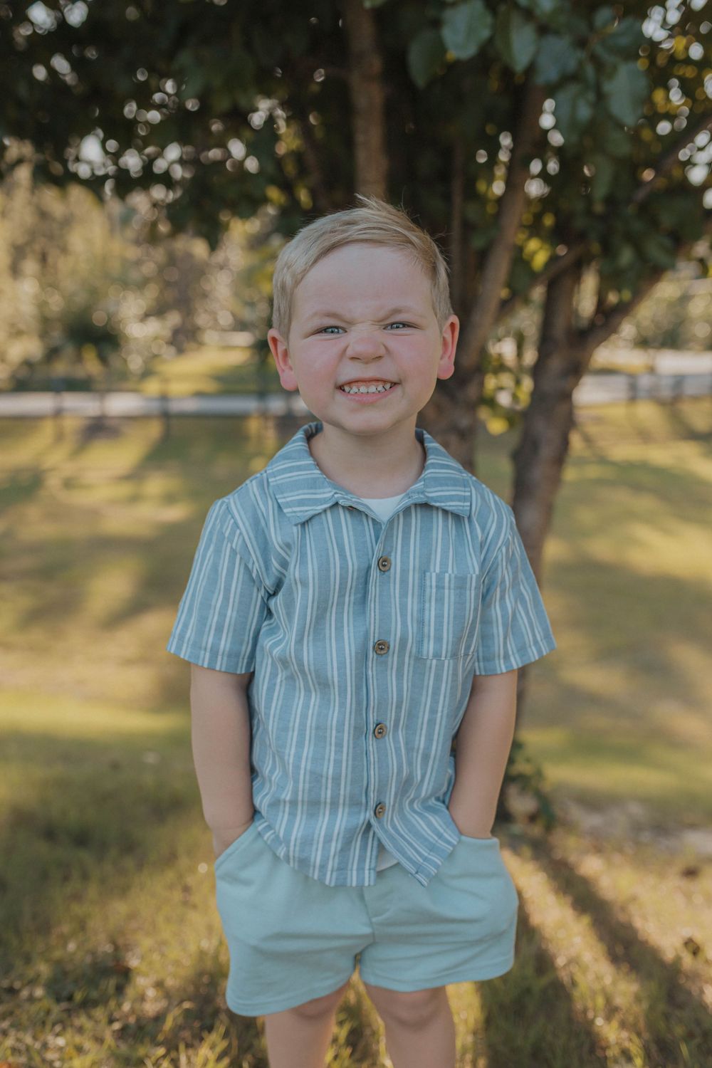 A child in a blue striped shirt and shorts poses outdoors with hands in pockets.