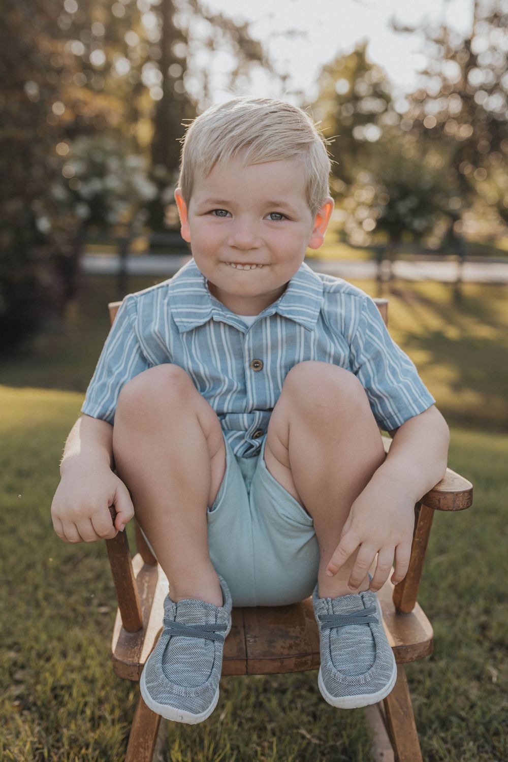 A child in striped clothing sits on a wooden chair outdoors with legs crossed.