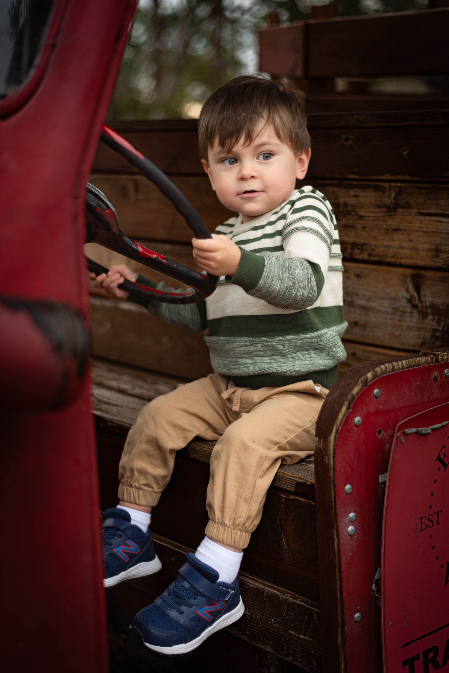 A young child sits in a vintage red theater seat wearing khaki pants, a striped shirt and blue shoes.