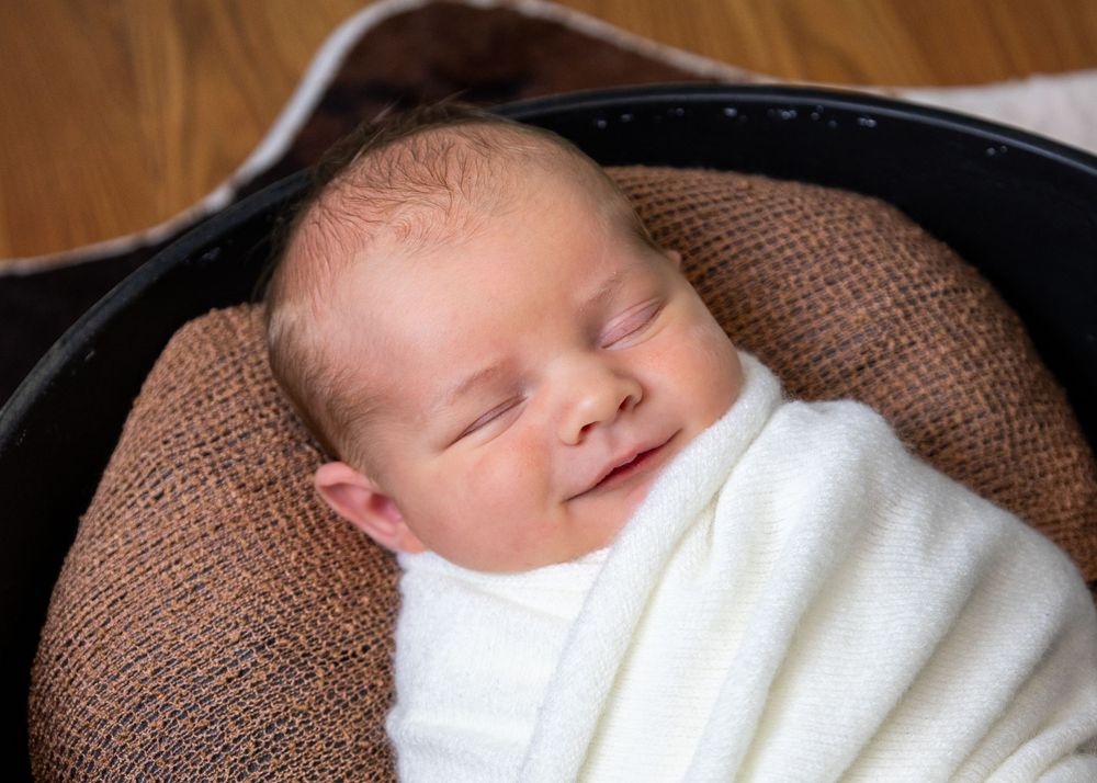 Sleeping baby wrapped in white blanket nestled in dark bowl with brown cushioning.