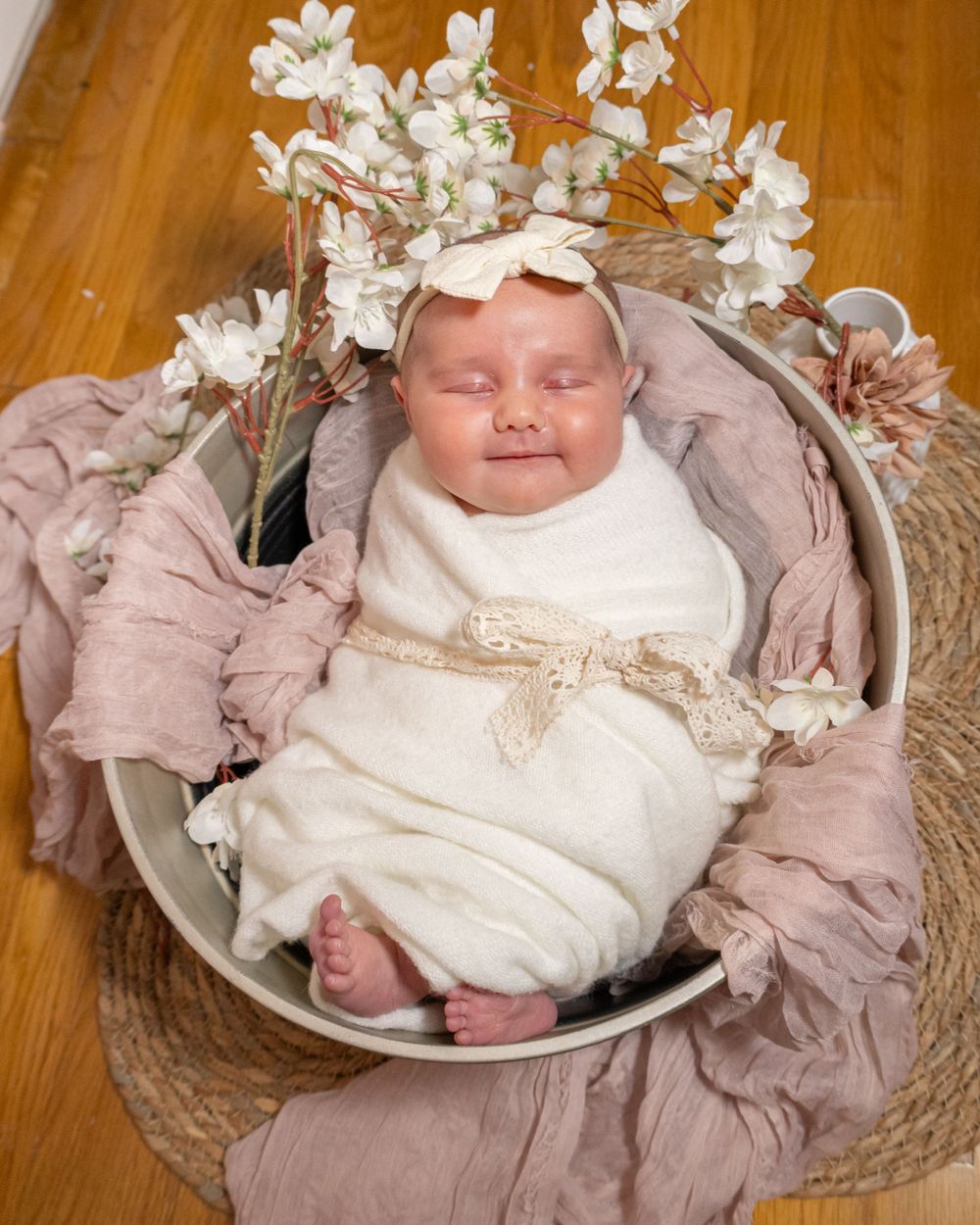 Sleeping baby wearing white dress surrounded by floral decorations on wooden floor.