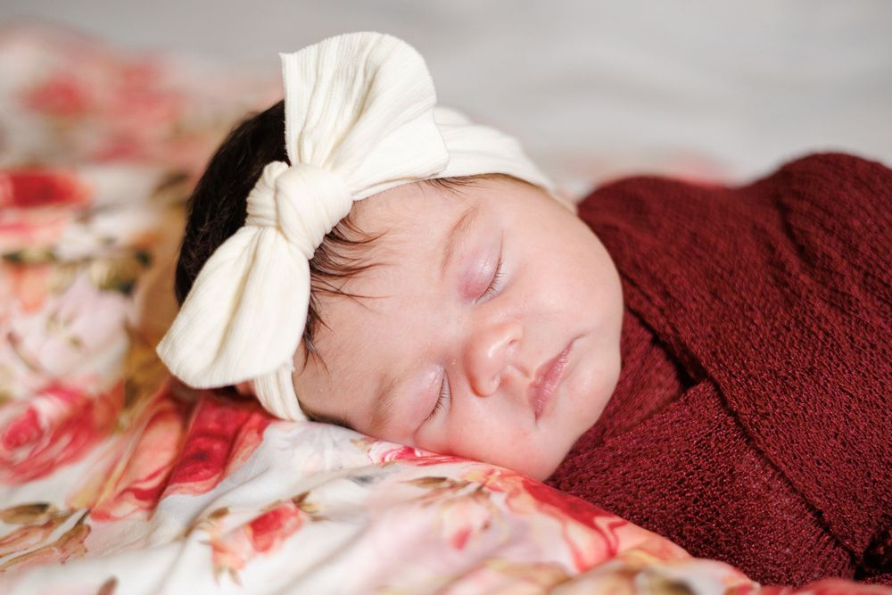 A sleeping newborn wearing a white bow headband wrapped in burgundy fabric.