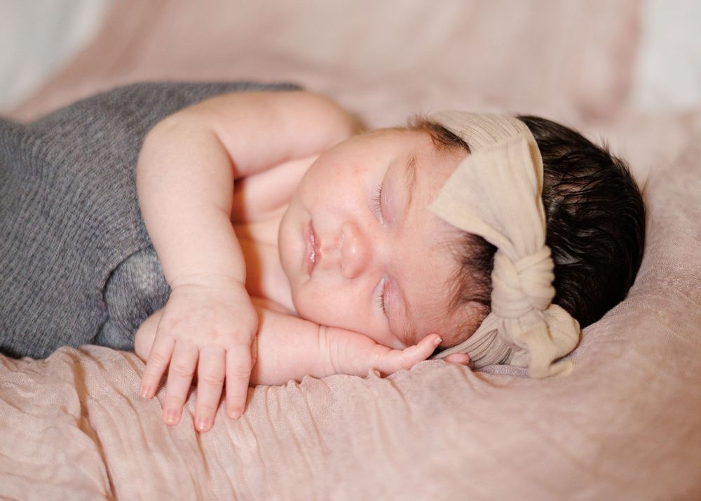 A peaceful newborn sleeping while wearing a cream bow and grey outfit.