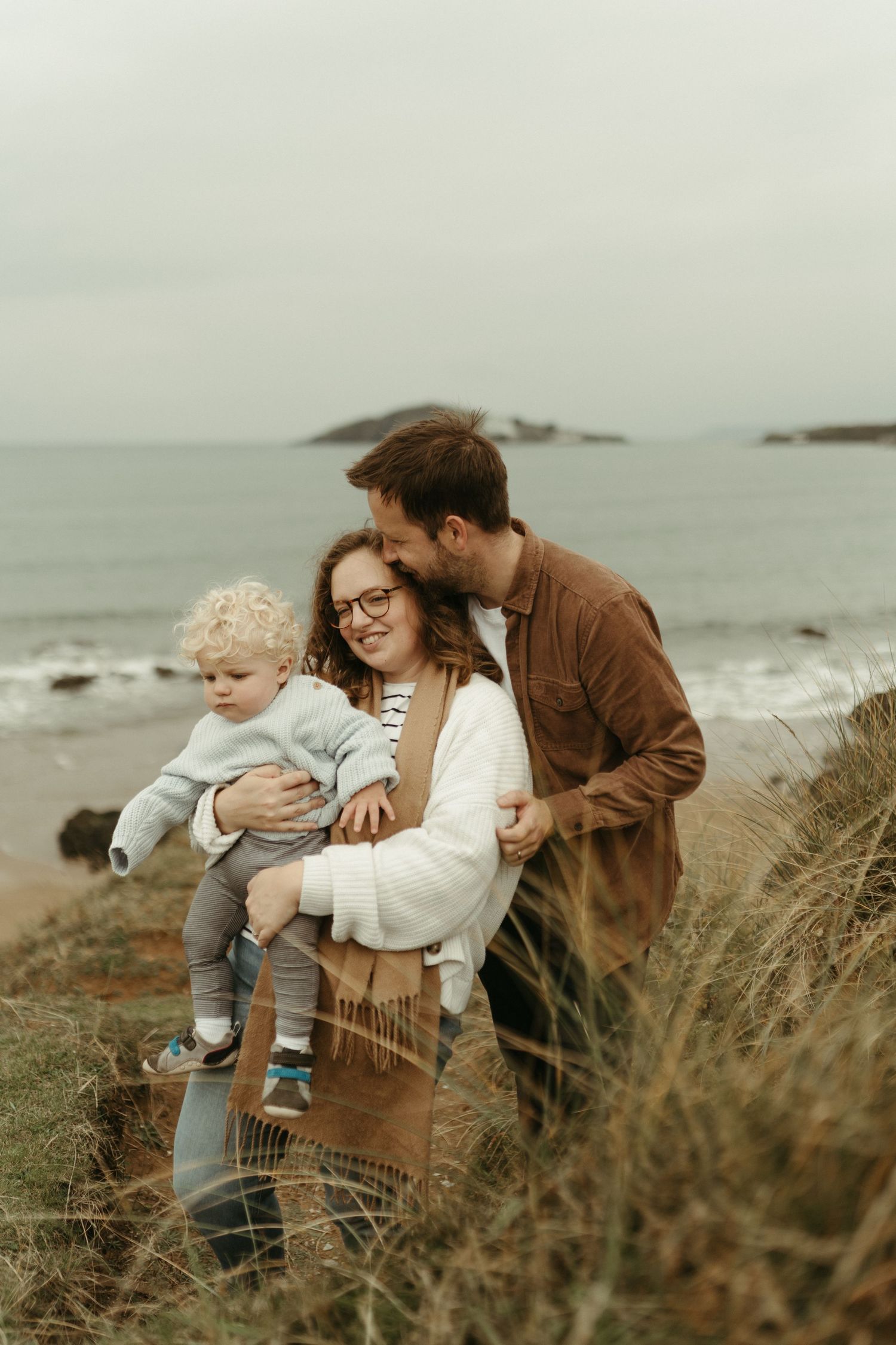Sweet family portraits at a misty coastal beach with tall grass and rocky shoreline in the background.