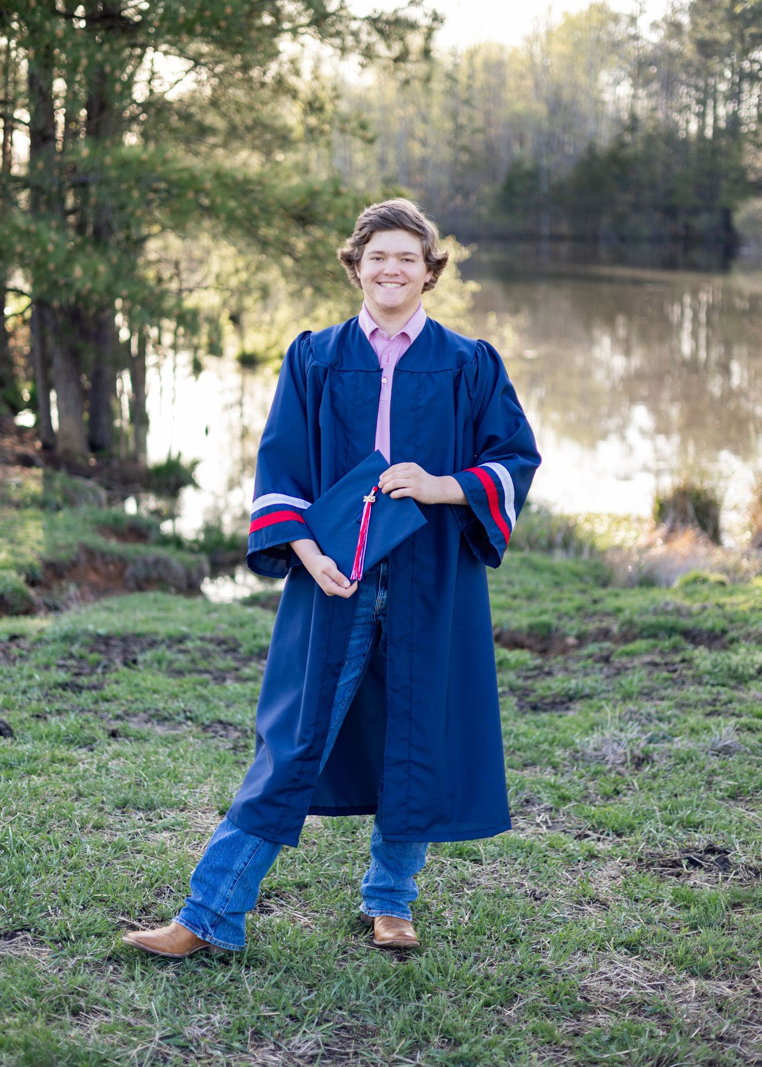 Graduate in navy blue gown posing on grassy hill near water.