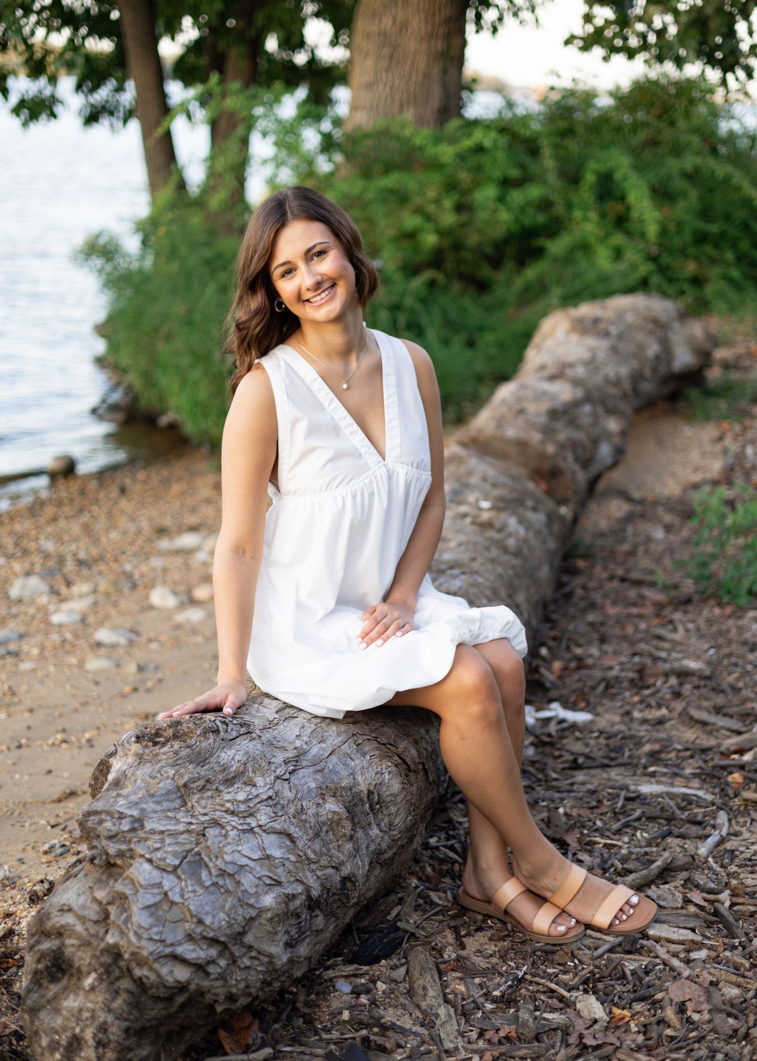 Model in white dress sits casually on fallen log by water's edge.