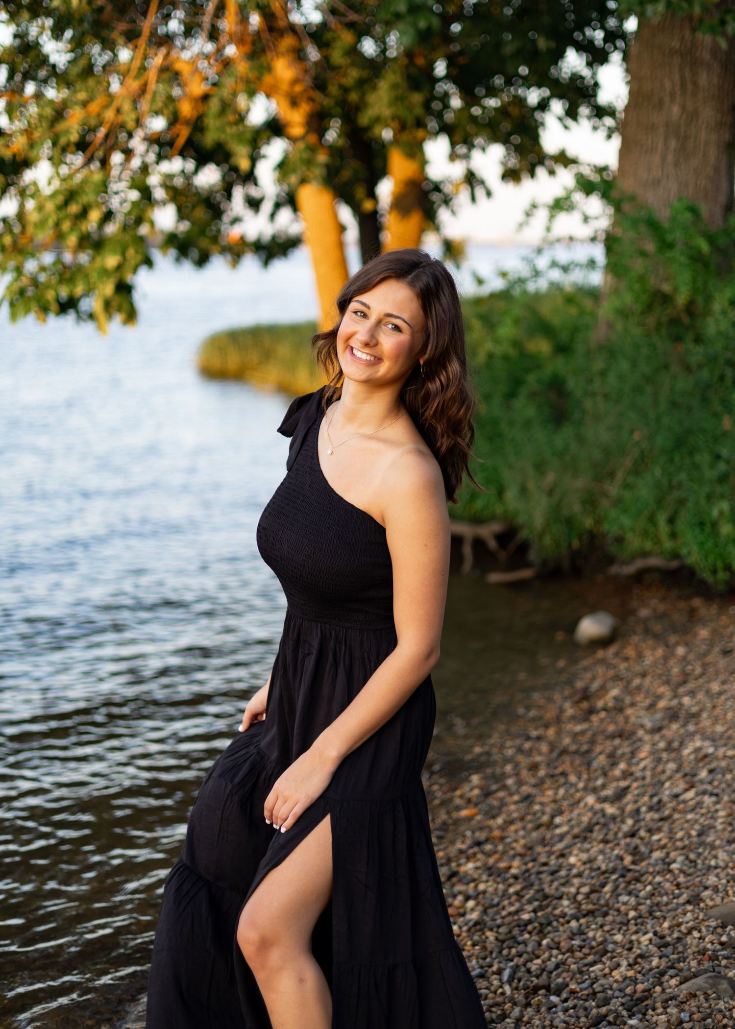 Person in black dress poses along rocky shoreline at sunset.