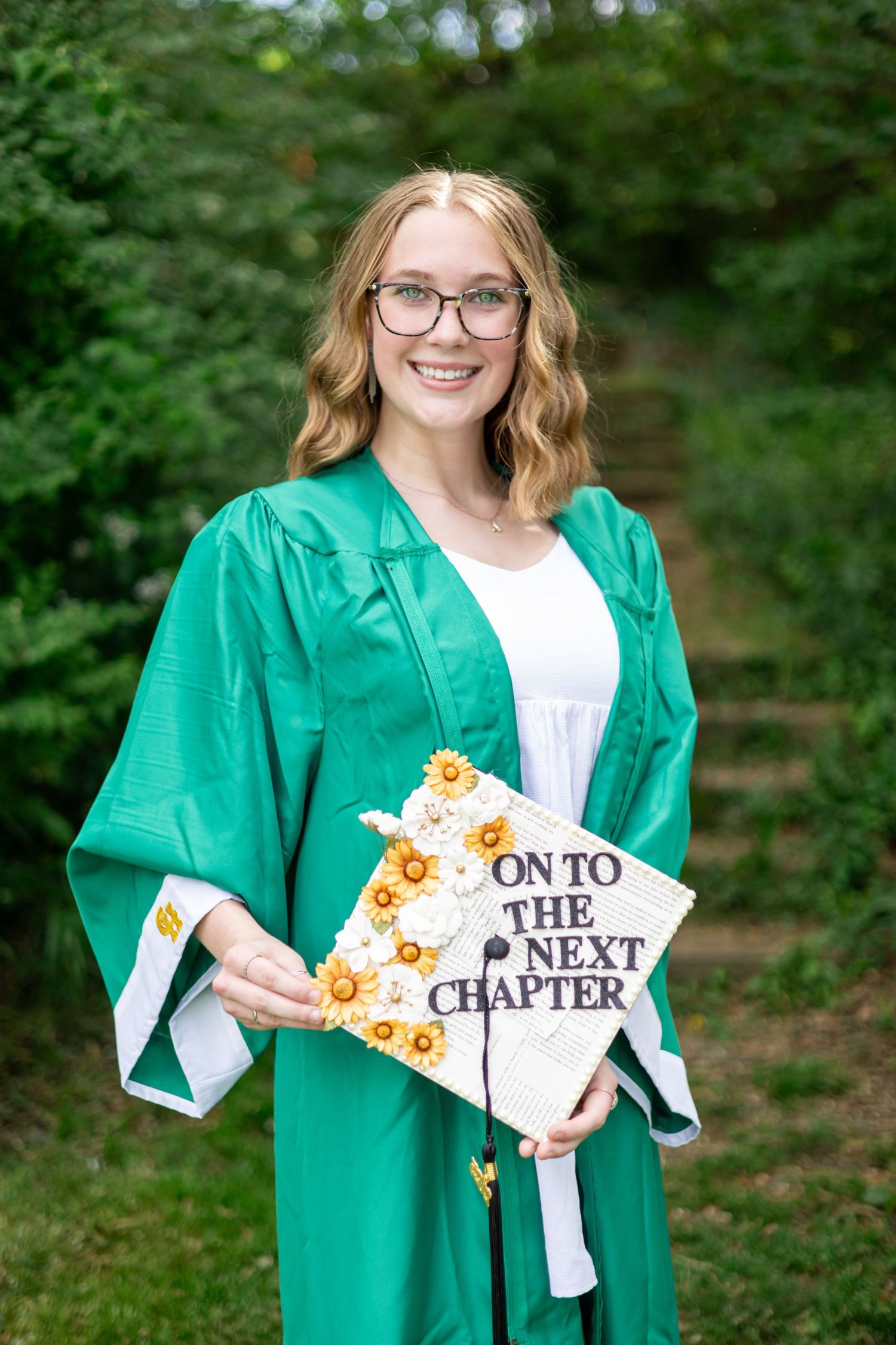 Graduate in green gown holding decorated cap with 'On to the Next Chapter' design.
