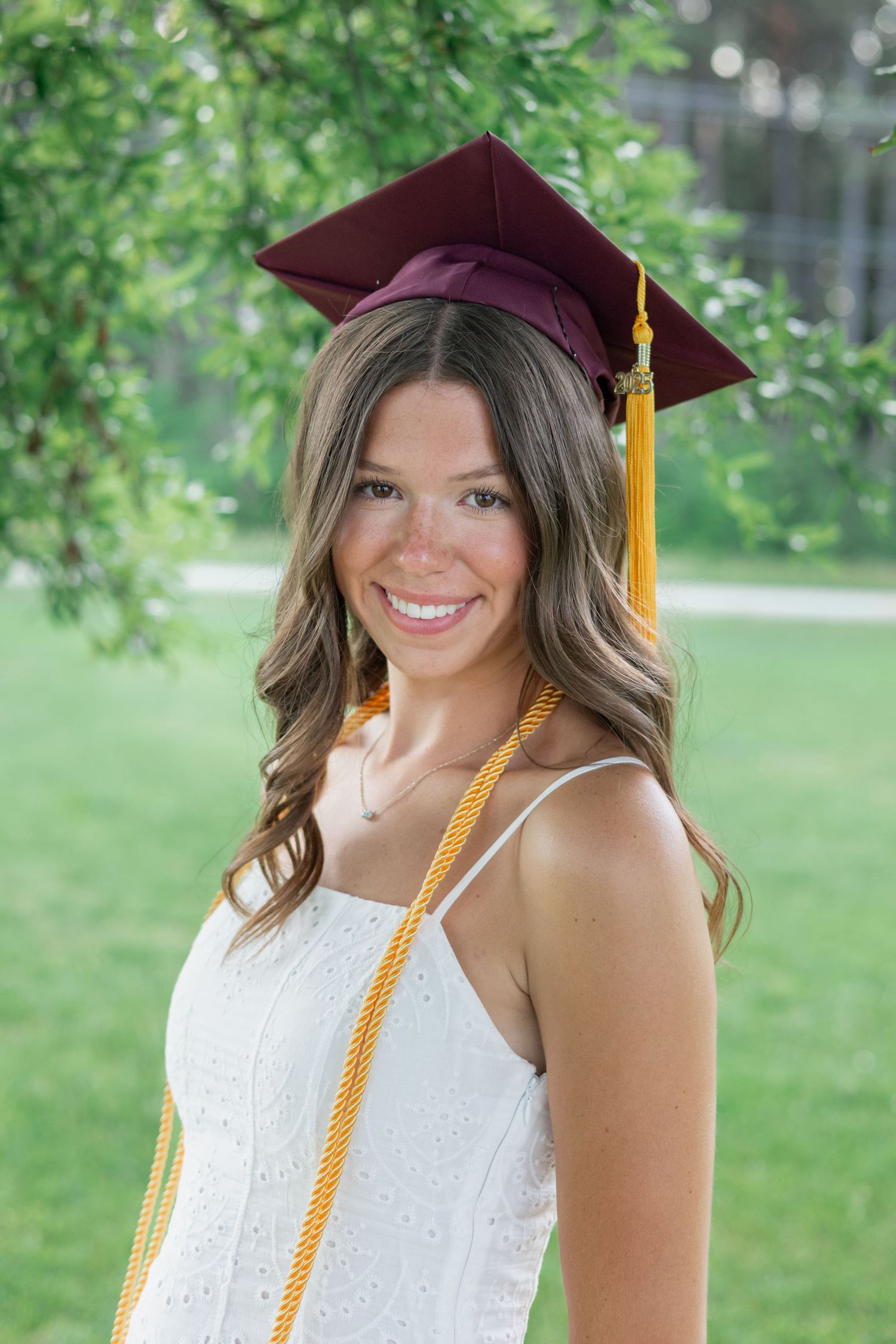 A graduate in maroon cap with gold tassel poses outdoors in white dress.