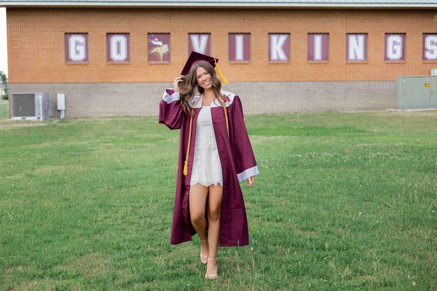 Graduate posing in a maroon cap and gown outside a school building during a sunny day.