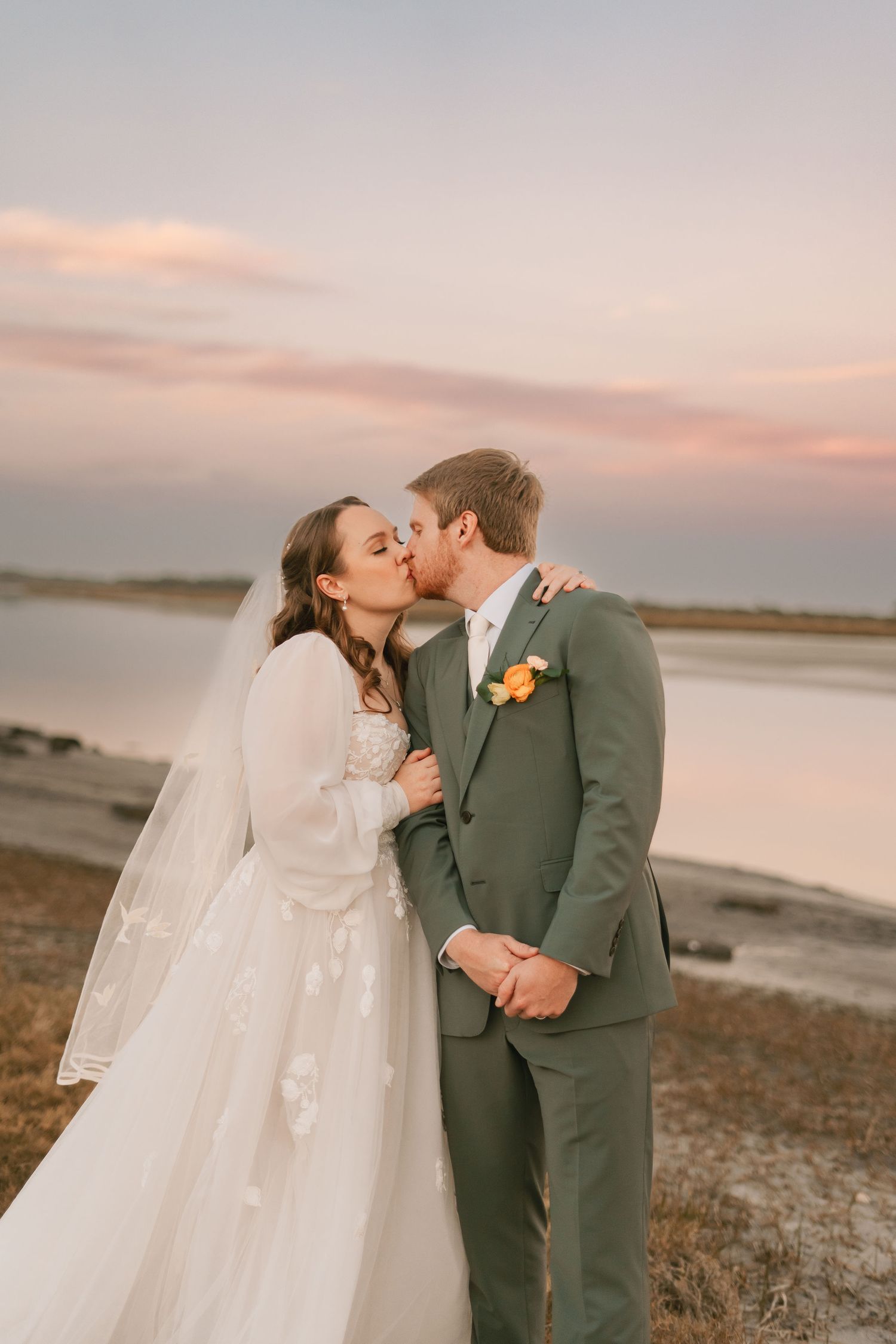 A romantic sunset wedding photo showing a couple sharing a kiss in a grassy field with a colorful floral bouquet.