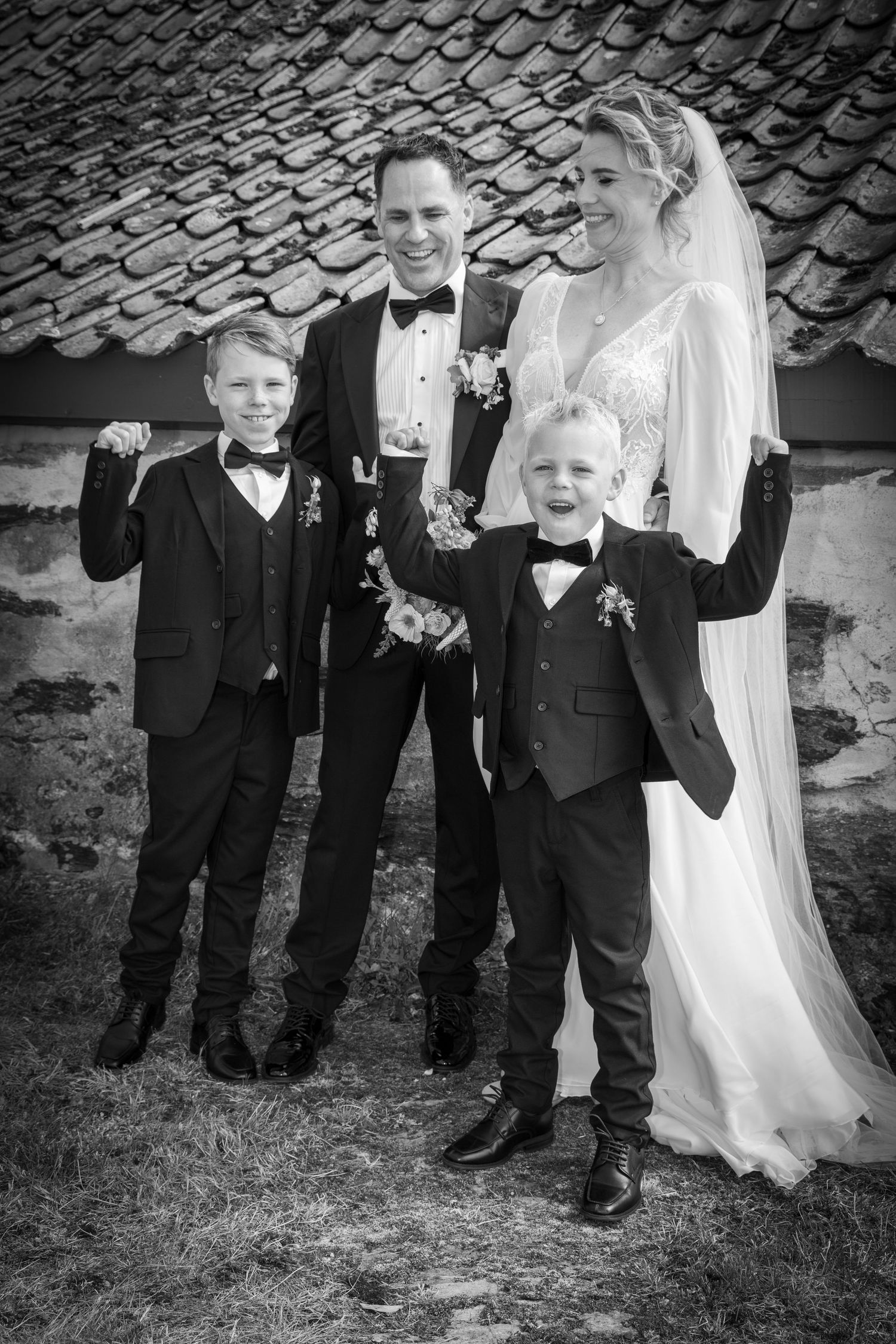 Joyful wedding portrait of bride, groom and two young boys in suits celebrating with raised fists.