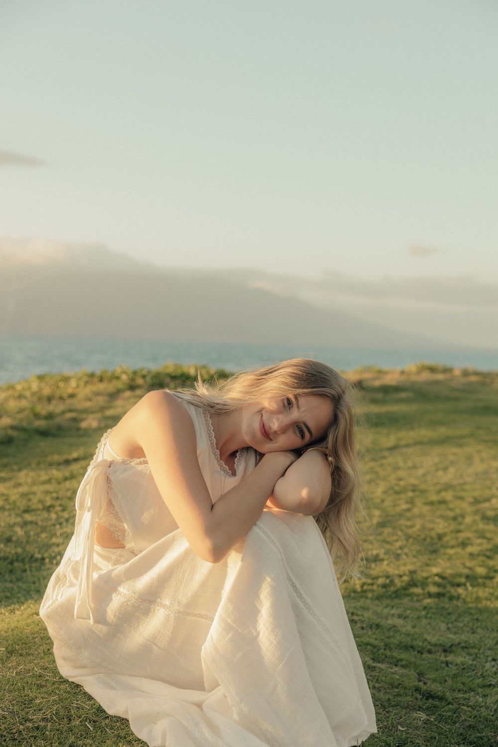 Person in a flowing white dress poses gracefully against a scenic green hillside landscape at golden hour.