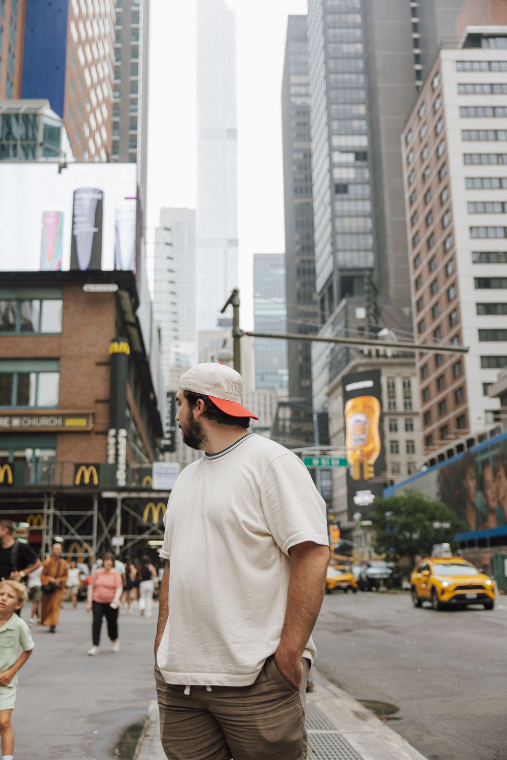 A person in a baseball cap stands on a busy New York City street corner with tall buildings and a yellow taxi in view.