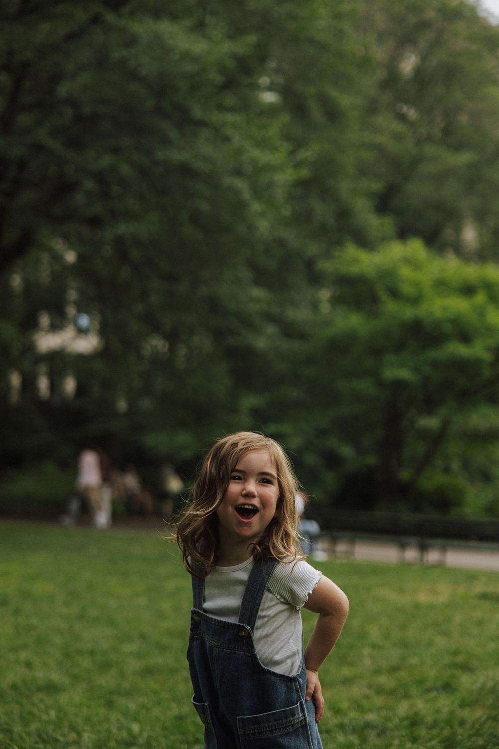 A child in denim overalls playing outdoors with a joyful expression in a green park setting.