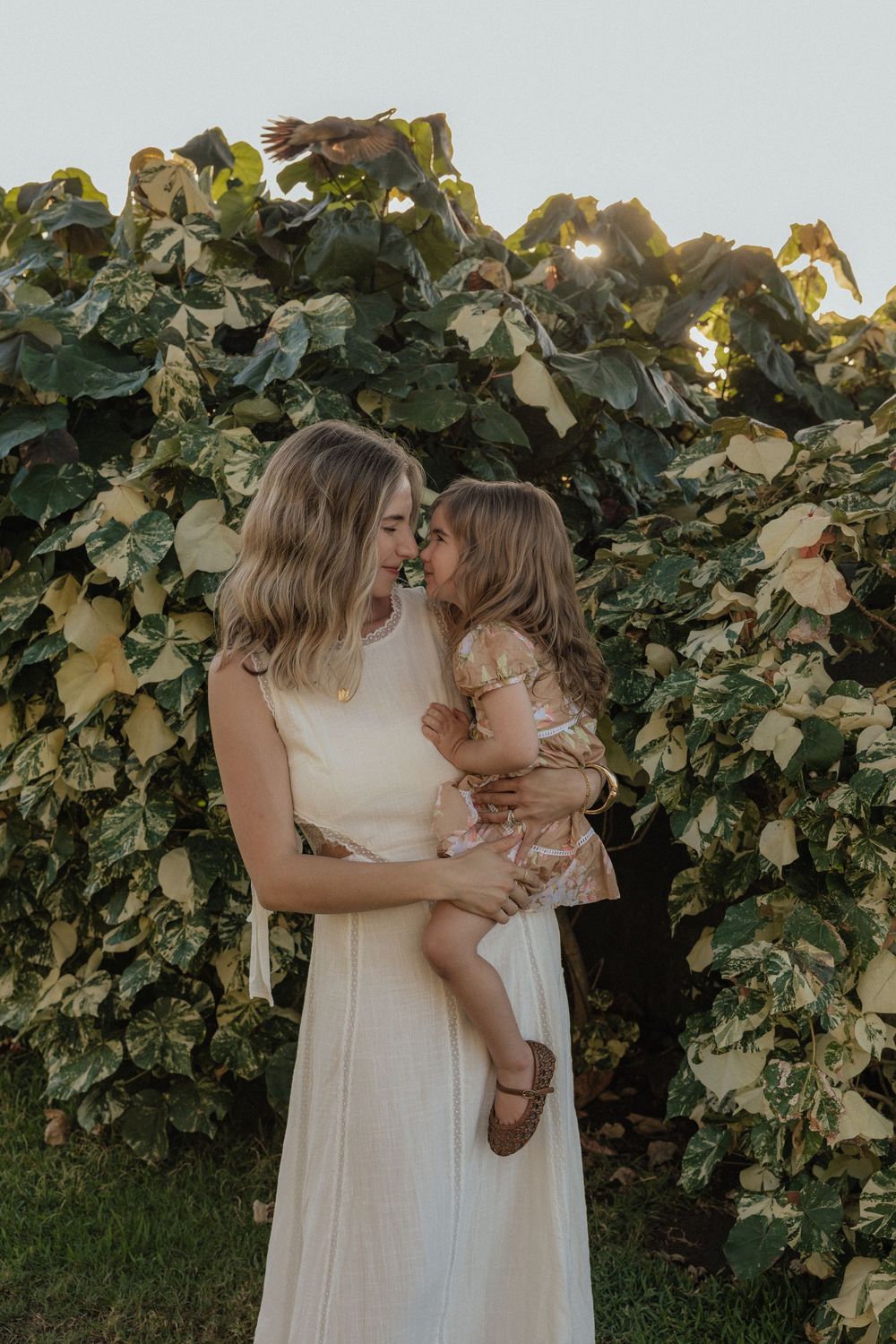 A tender family moment captured in a white dress against lush green foliage at sunset.