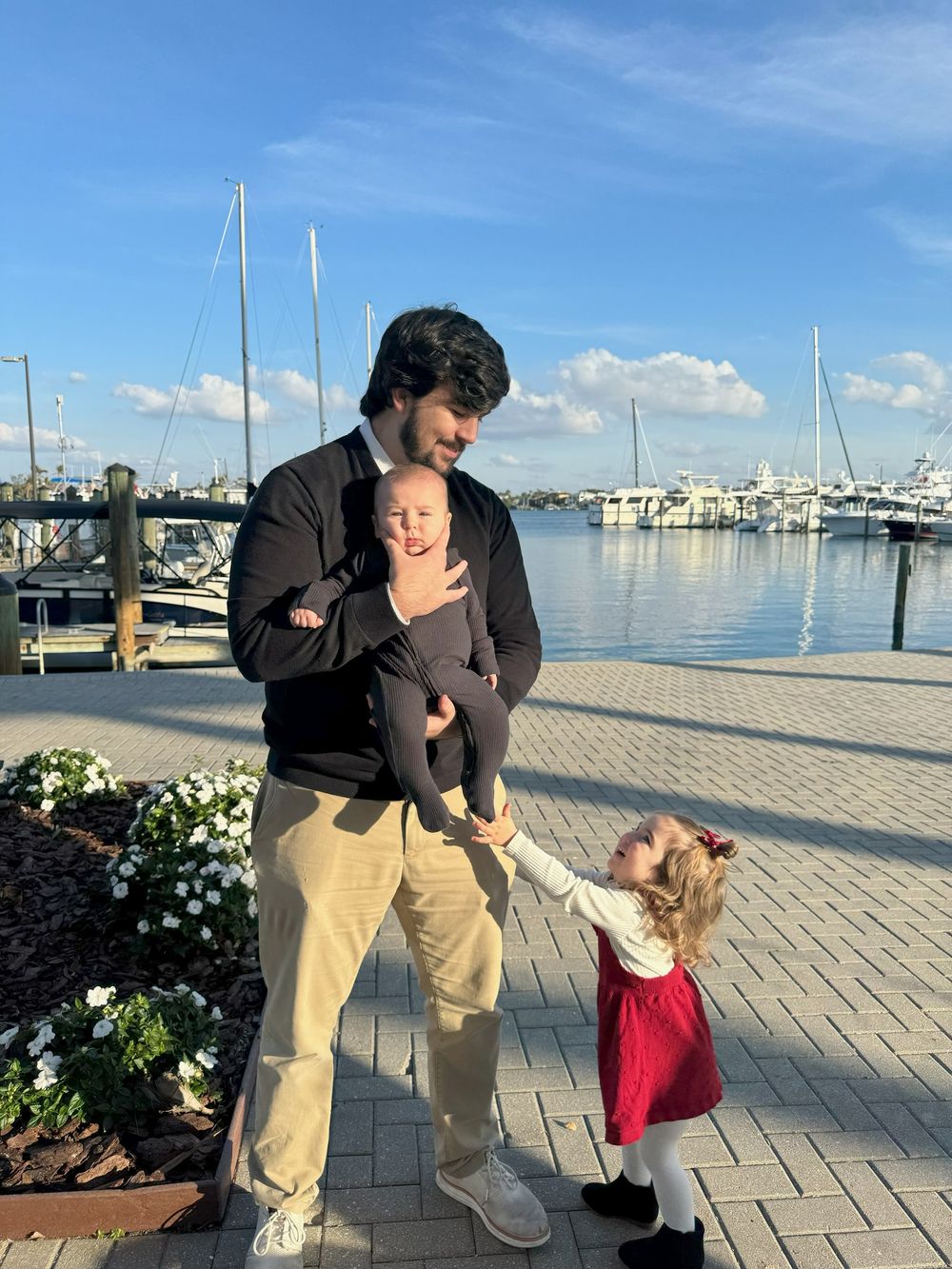 A family enjoys a sunny day at a marina with boats docked in the background and flower planters along the walkway.