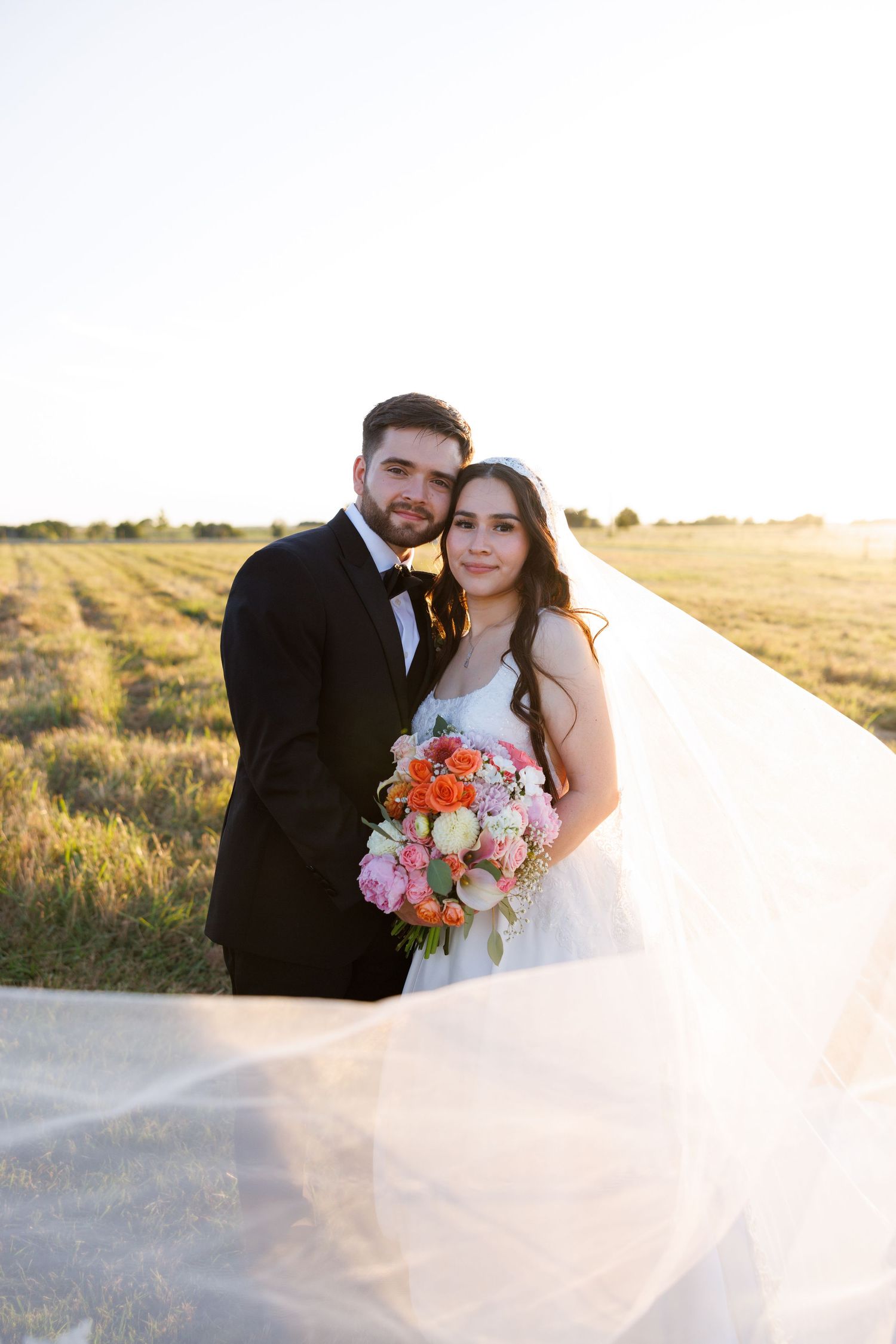 A romantic sunset wedding portrait in a golden field with a flowing veil and colorful bouquet.