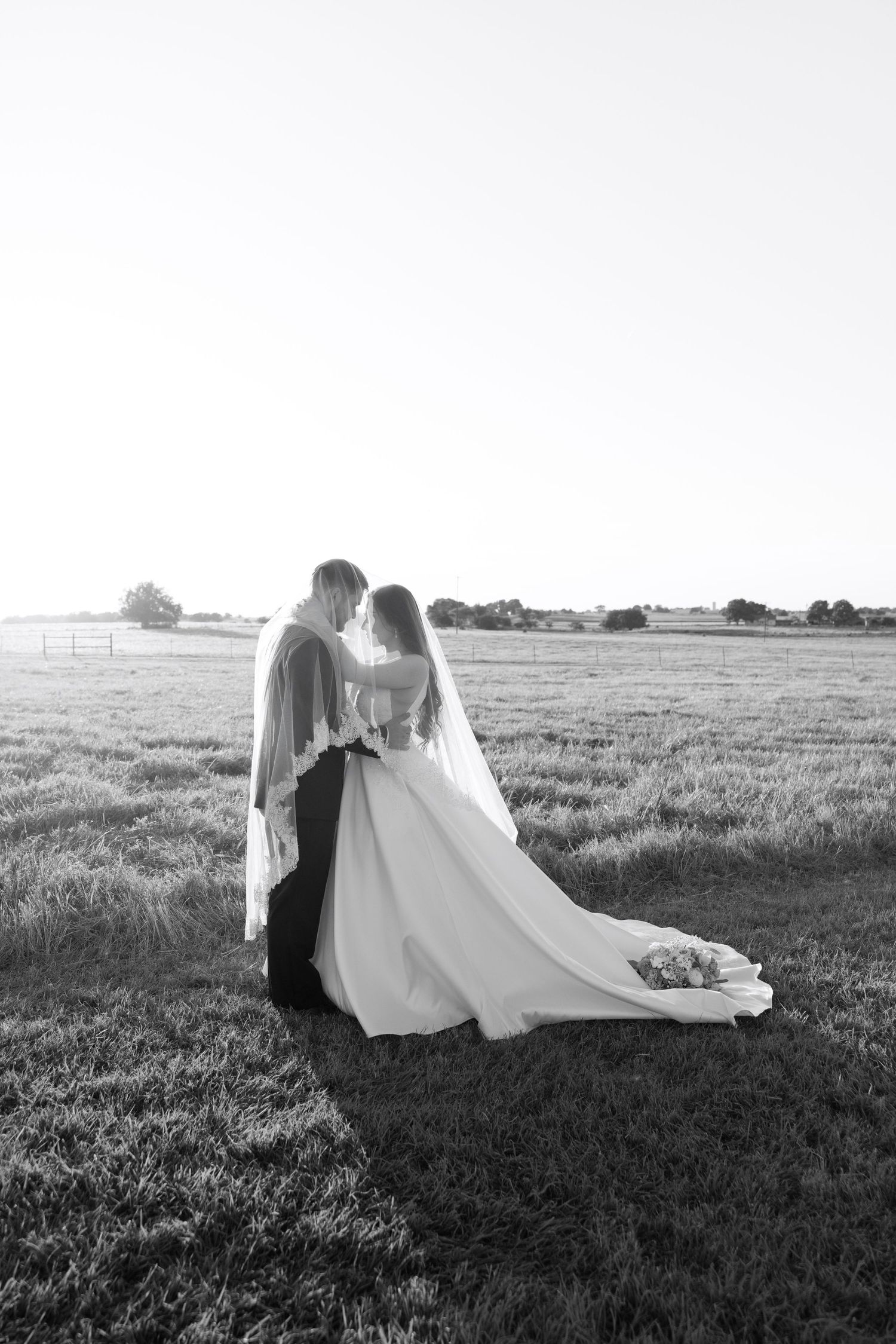 A wedding couple poses in an open field with flowing veil and dramatic lighting.