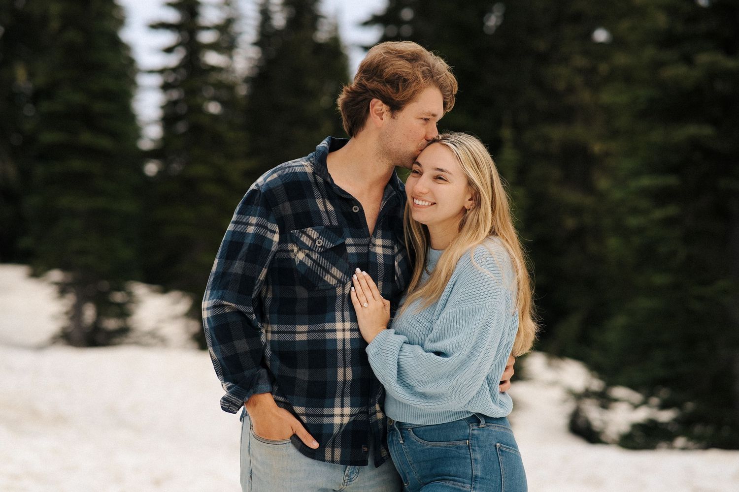 A couple in casual winter clothing shares an intimate moment in the snow with evergreen trees in the background.