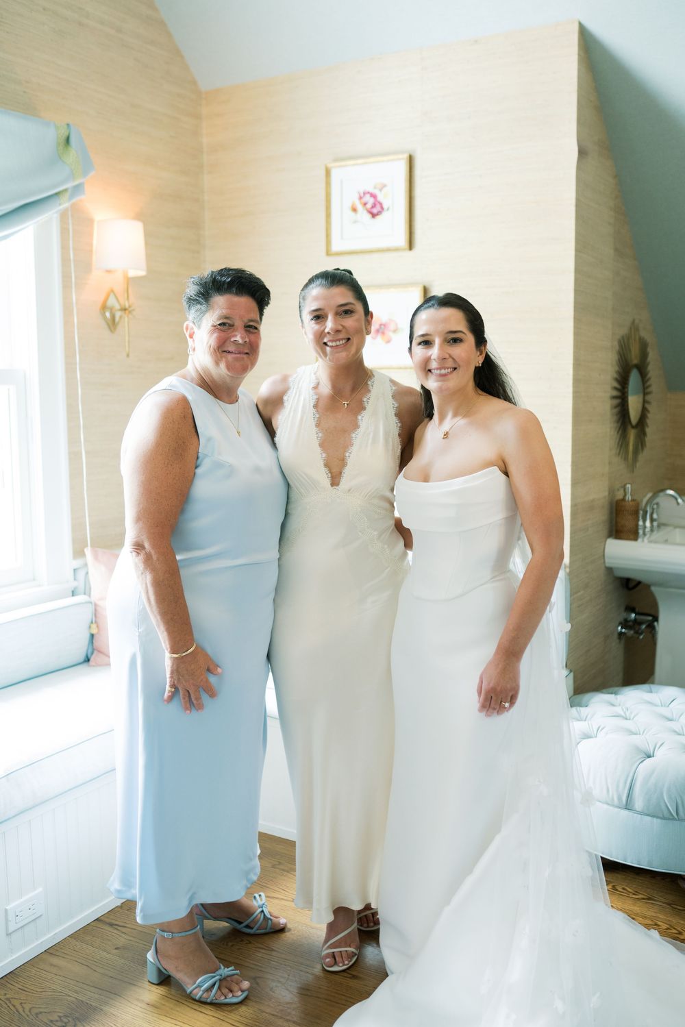 Wedding party members in white and light blue dresses pose together in a bright room.