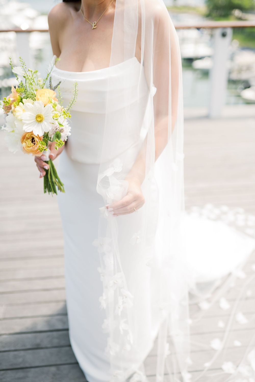 Close-up details of a bride's white wedding dress and pastel bouquet on a sunny deck.