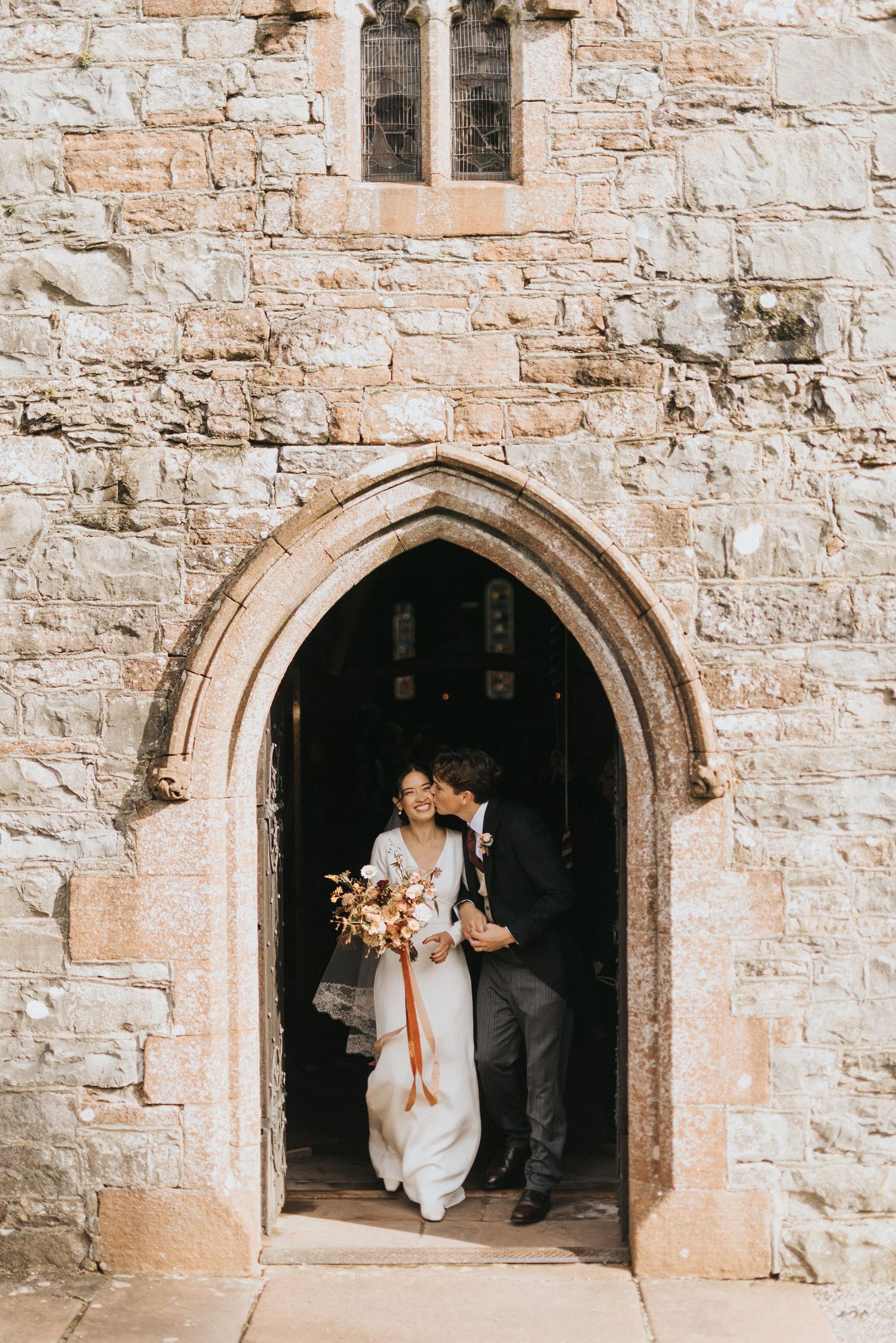 Groom kisses bride as they exit a historic stone church doorway with Gothic arched entrance and rustic textured walls at Askham Hall.