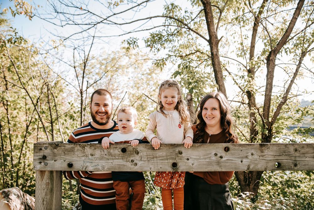 A family poses together on a rustic wooden bridge surrounded by trees on a sunny day.