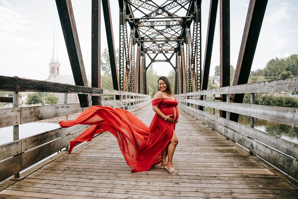 A flowing red dress catches the wind on a wooden bridge with metal trusses at golden hour.