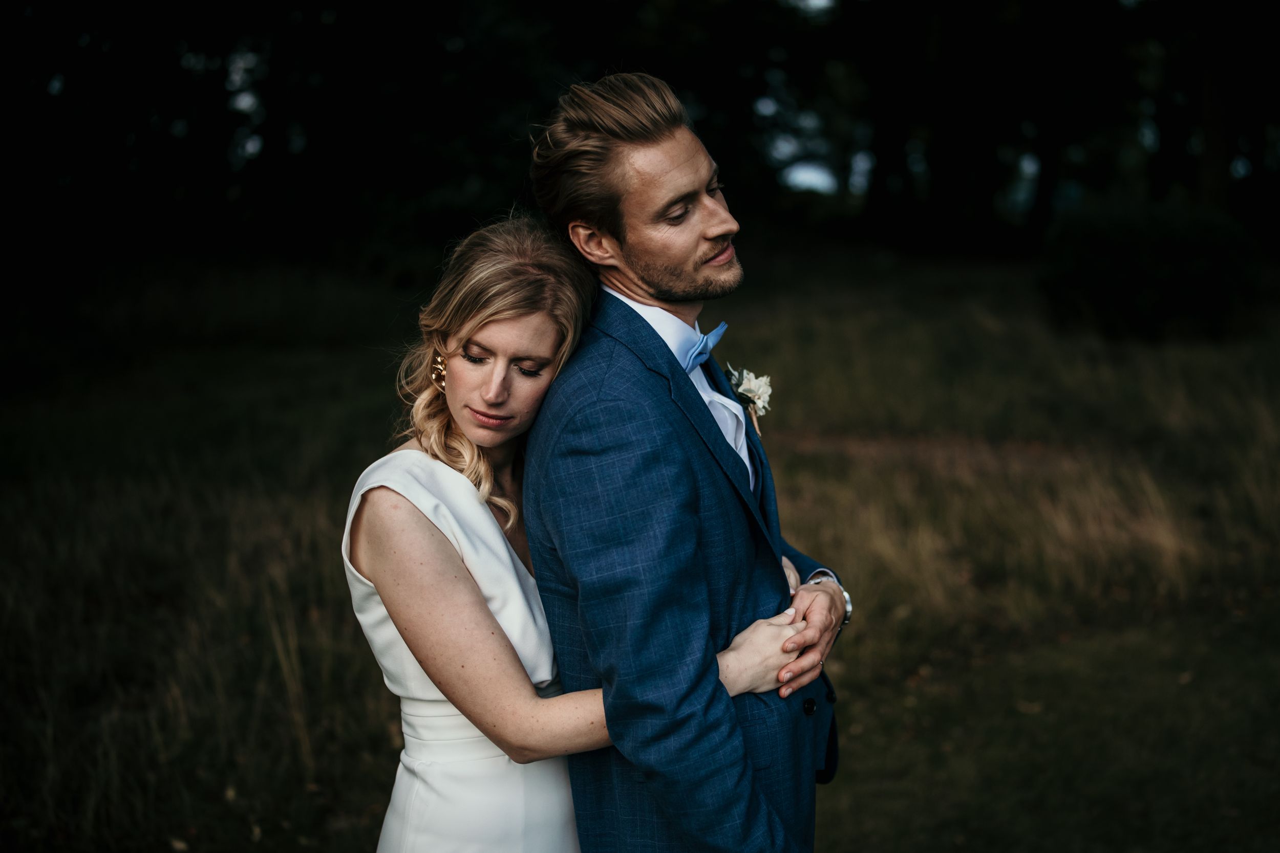 A couple poses together in a moody outdoor setting wearing formal attire.