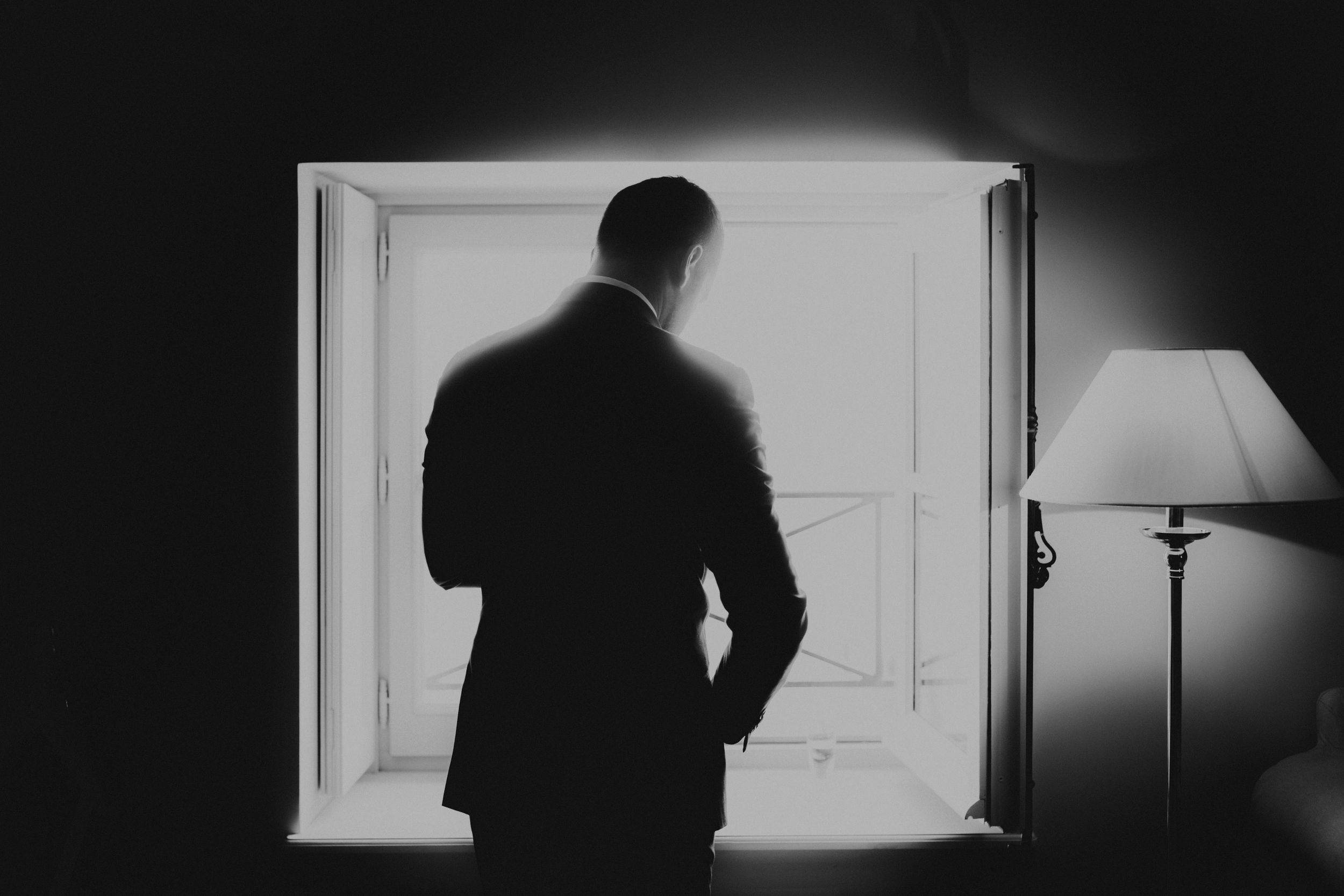 A silhouette of a figure stands by a window next to a table lamp in dark monochromatic lighting.