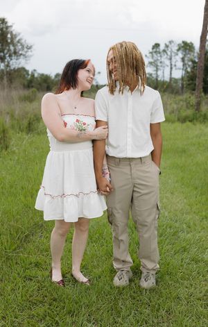 A couple in white and khaki outfits standing together in a grassy field on a sunny day.