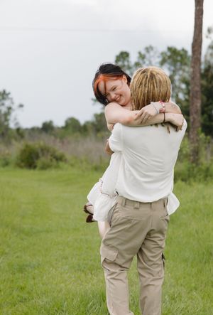 Playful couple embracing and spinning in a green meadow with pine trees in the background.