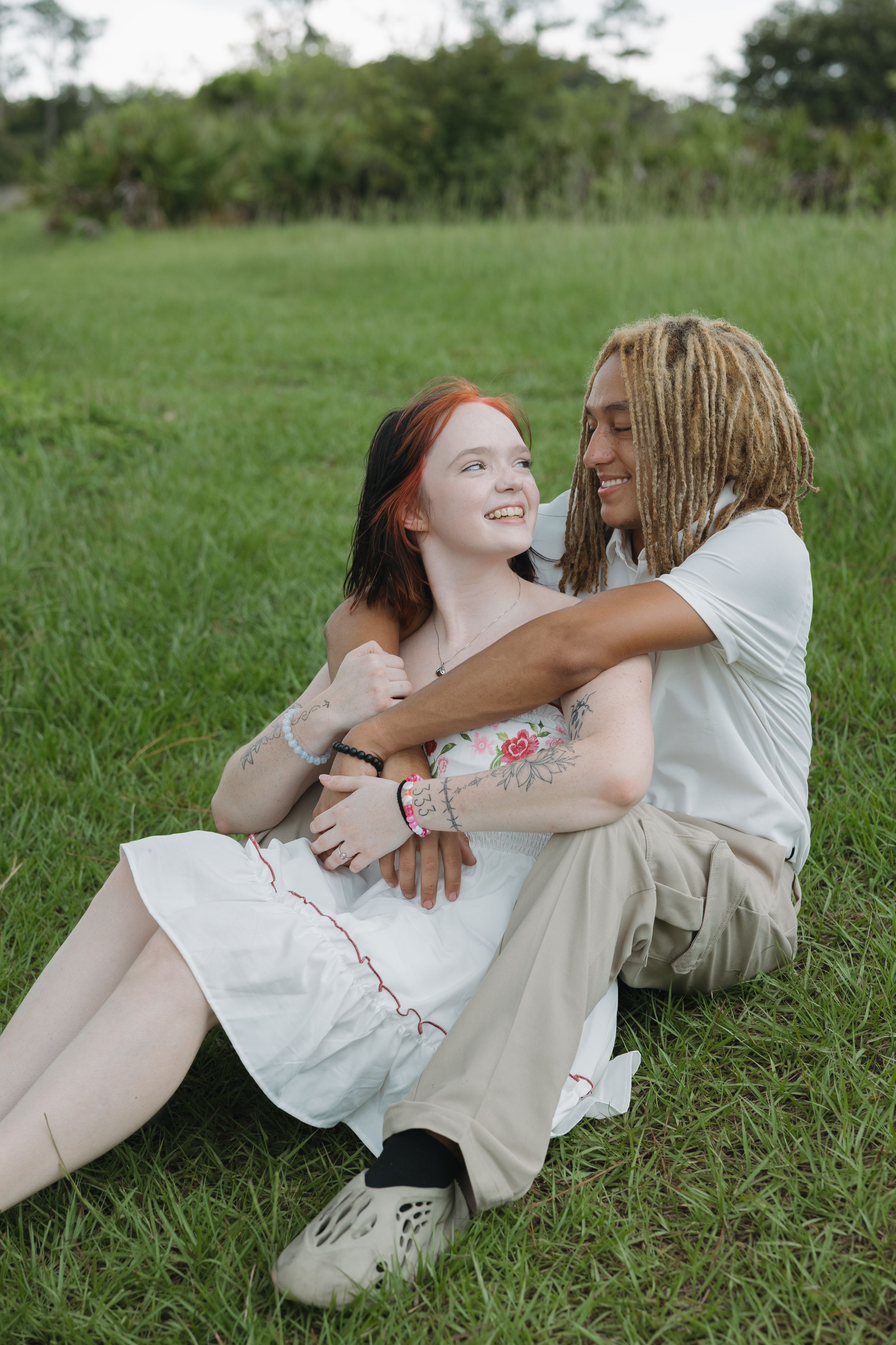 Two people in light colored dresses playfully lying in green grass during a sunny day.
