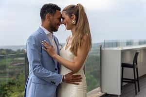 Couple in light blue suit and white dress share an intimate moment on a balcony overlooking greenery.