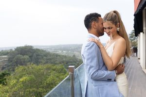 Couple embraces on balcony with scenic forest view, bride in white dress and groom in powder blue suit.