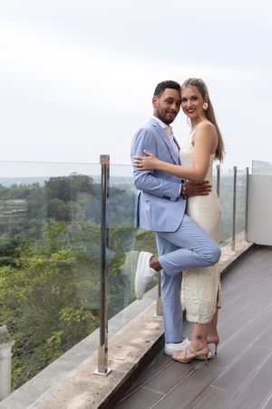 Romantic couple pose on scenic balcony, with bride in lace dress and groom in light blue suit.