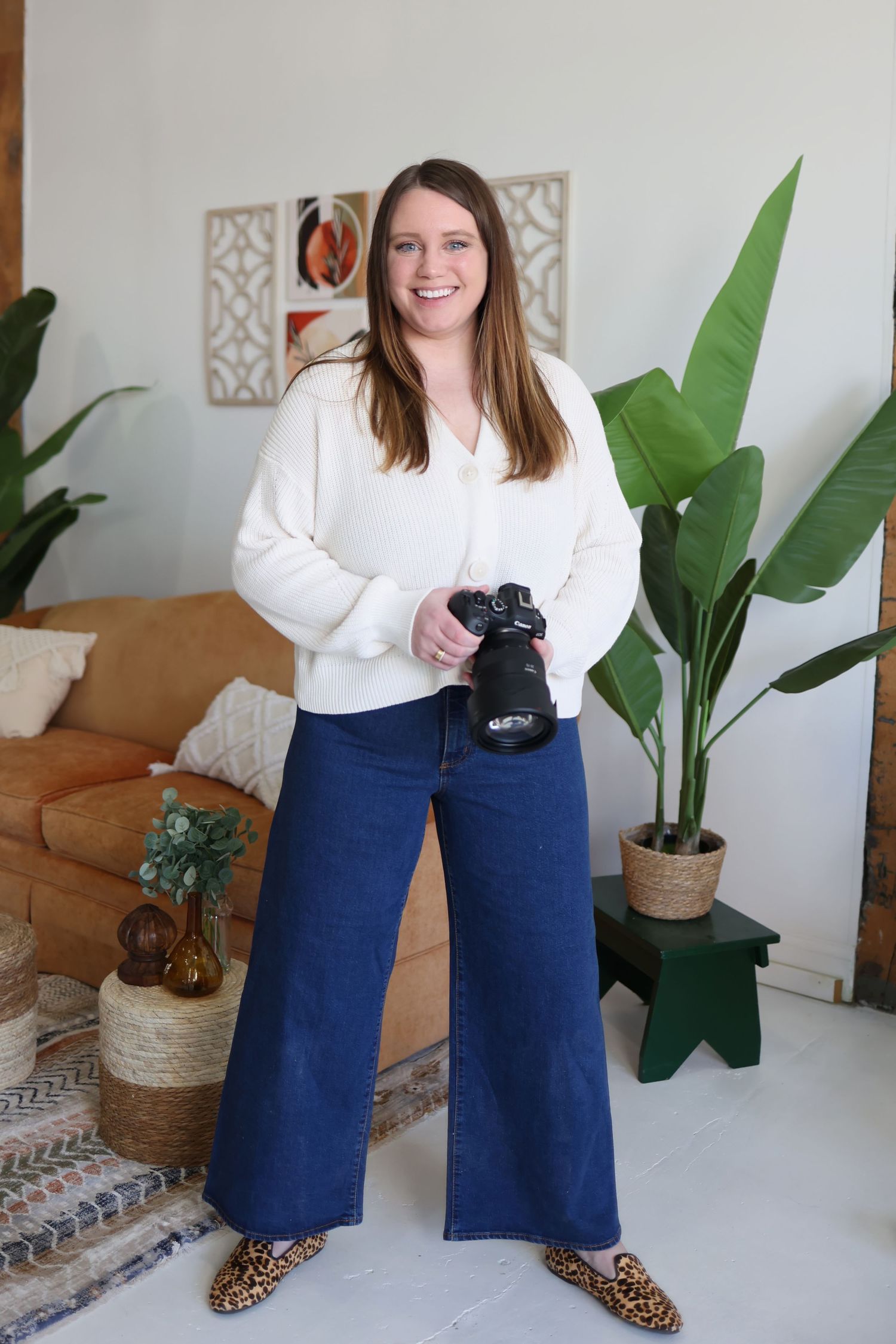 Professional photographer in white sweater and jeans poses with camera in modern living room setting.